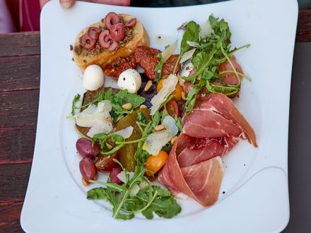 Auf dem Bild ist ein leckerer Antipasti-Teller mit Oliven, getrockneten Tomaten, Schinken, Rucola etc. zu sehen.